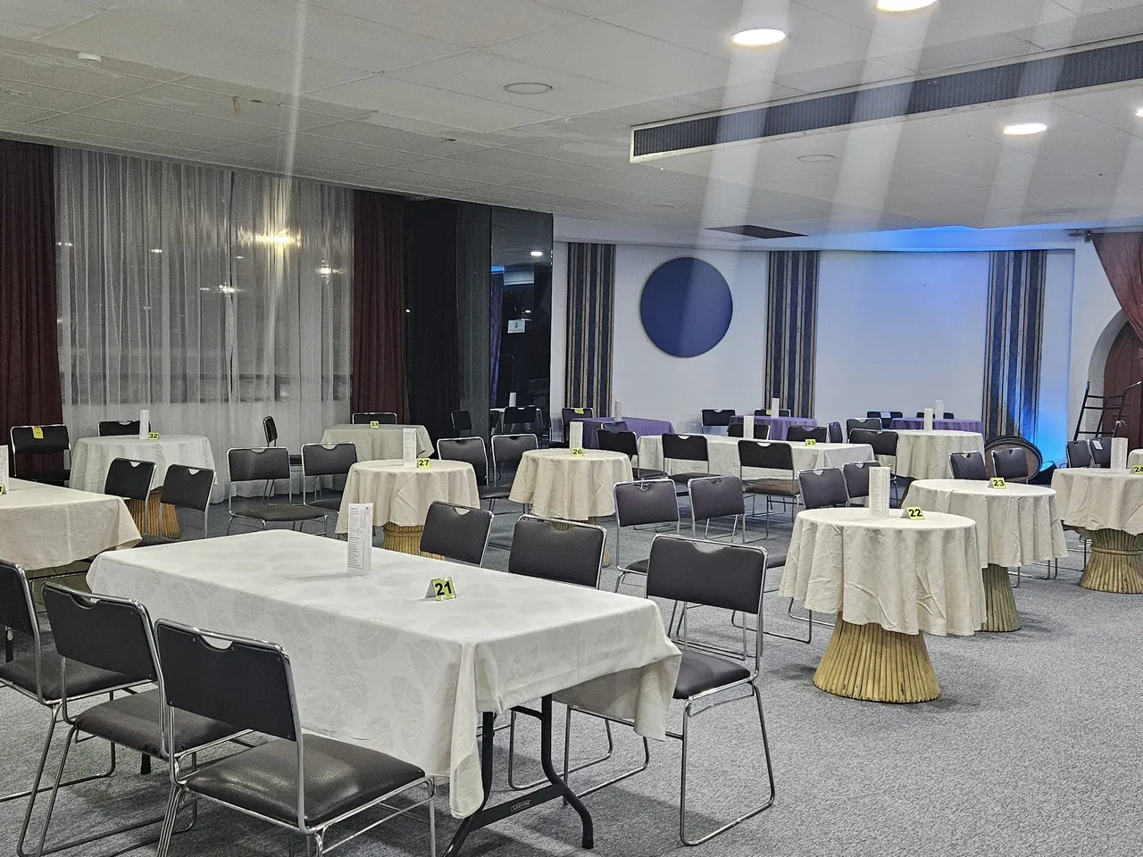 Salón del hotel montado para un banquete: mesas largas y redondas con mantel crema, sillas negras y el escenario iluminado al fondo