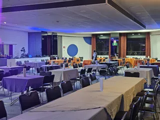 Salón montado: mesa larga vestida en primer plano y mesas redondas con mantel naranja al fondo, bajo luz azul