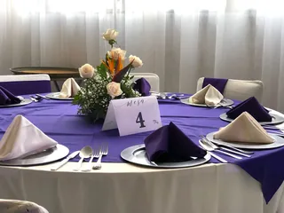 Mesa redonda vestida con mantel morado, servilletas dobladas en pico, cubiertos y un centro de rosas blancas y naranjas