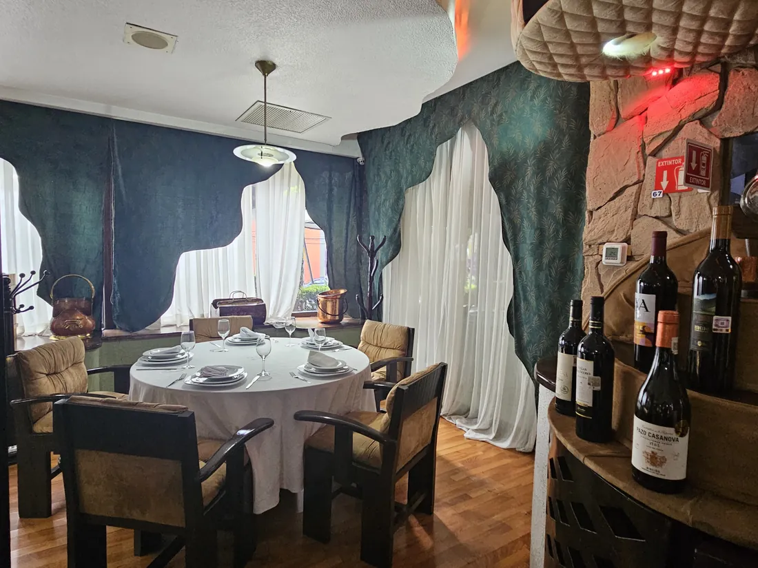 Mesa redonda de La Cava del Duque, vestida con mantel blanco, copas y servilletas dobladas, entre la cortina verde y la columna de piedra