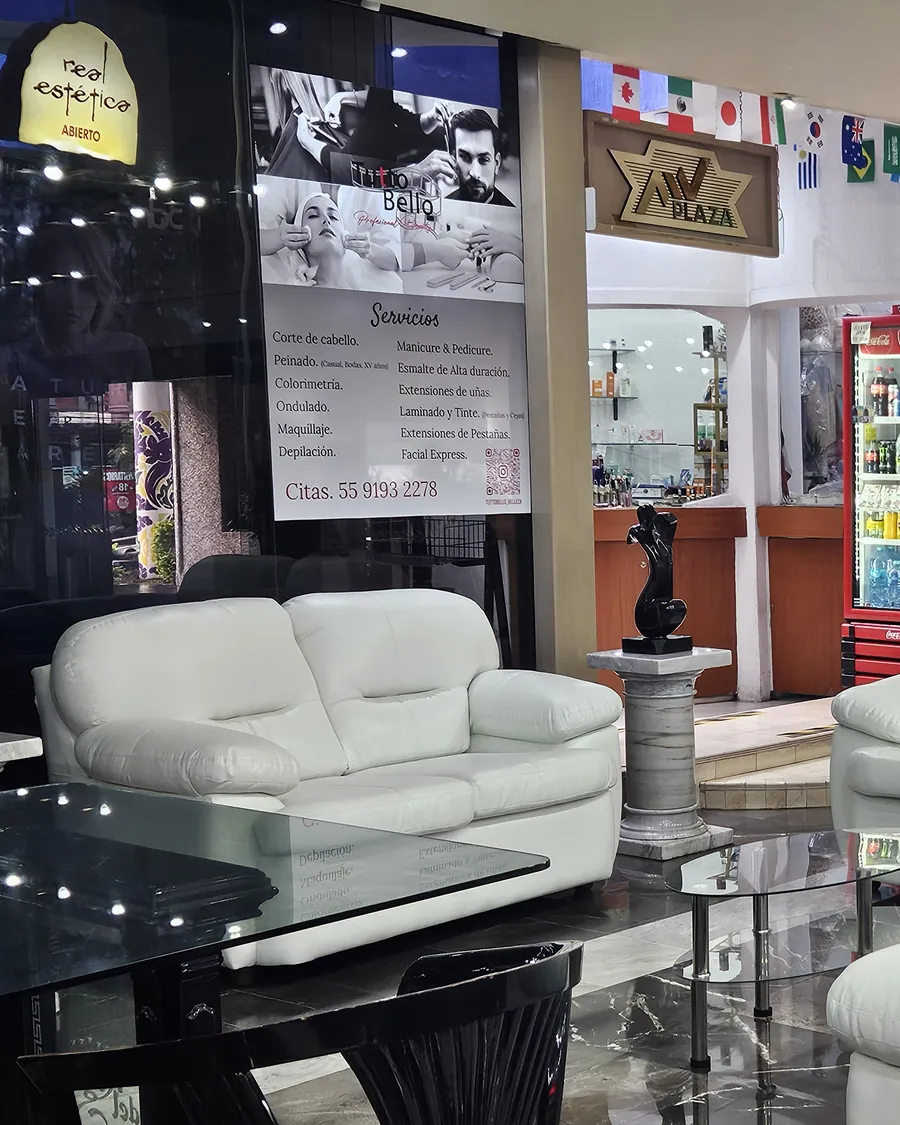 Escaparate de la estética en el vestíbulo del hotel: letrero luminoso encendido, el cartel de servicios de Tutto Bello y un sofá blanco delante